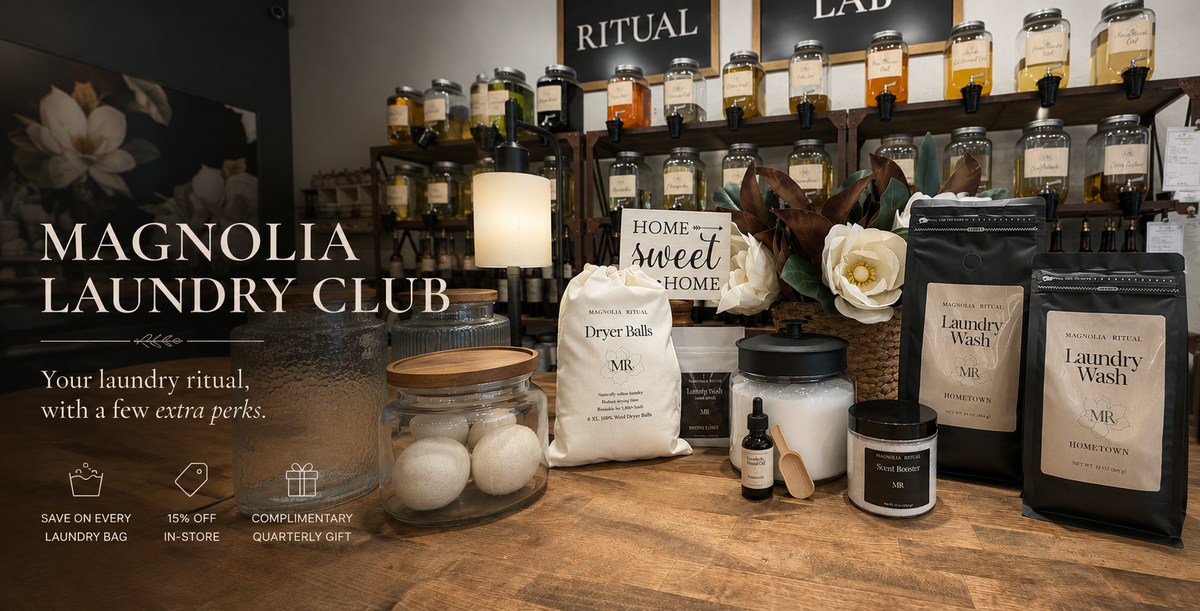 A styled product display for Magnolia Laundry Club featuring various laundry care products including bags of Laundry Wash, dryer balls in a glass jar, a candle, and other home fragrance items on a rustic wooden table, with shelves of similar products in the background.